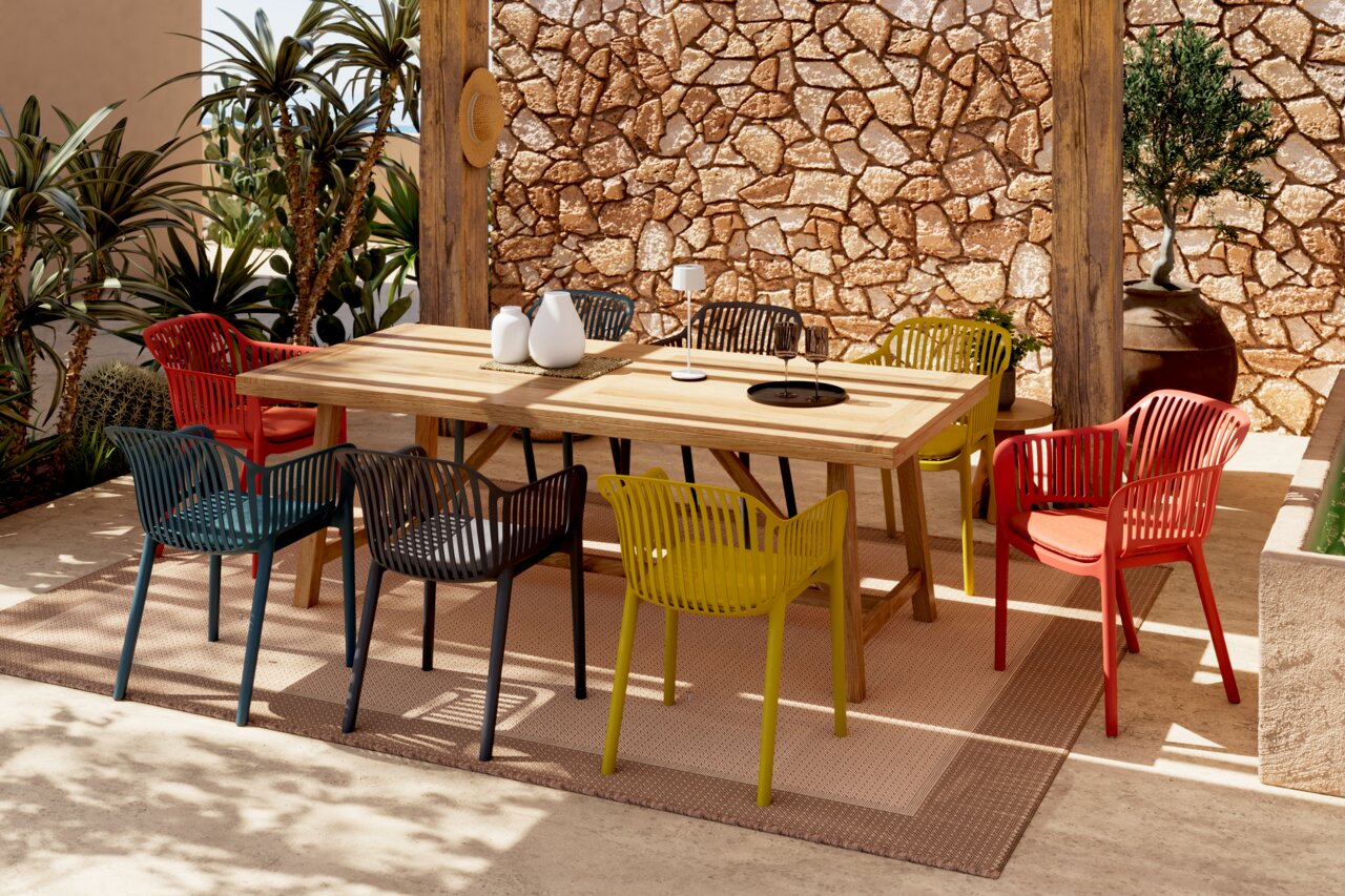 Kleurrijke stapelstoelen rond een houten tafel op een terras met een stenen muur en planten, gezien vanuit een zijdelingse hoek.