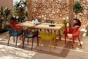 Kleurrijke stapelstoelen rond een houten tafel op een terras met een stenen muur en planten, gezien vanuit een zijdelingse hoek.
