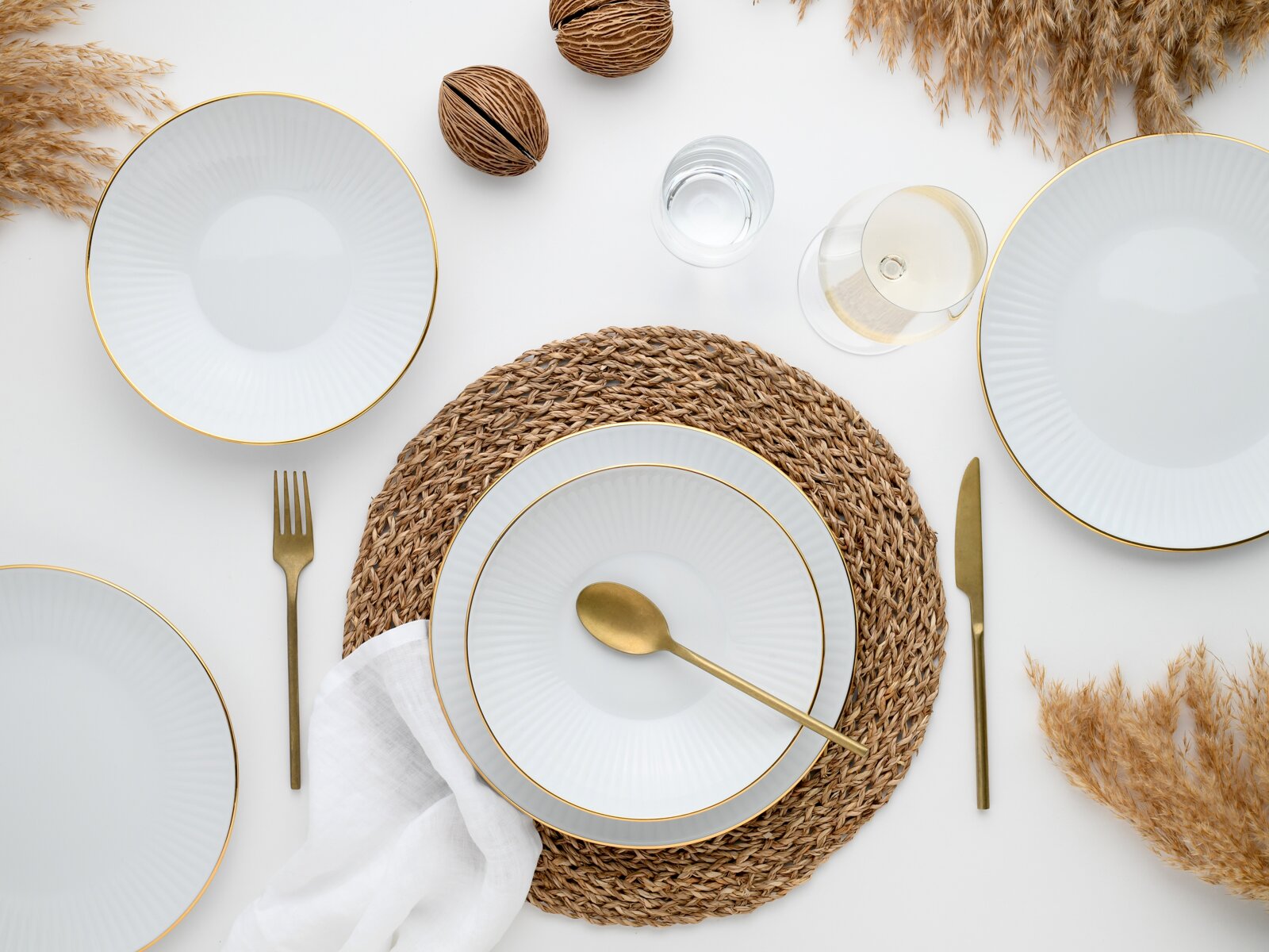 Draufsicht auf ein elegant gedecktes Tischarrangement mit weißen Tellern mit goldfarbenem Rand, goldfarbenem Besteck, geflochtenem Platzset, Weinglas und Wasserglas auf hellem Hintergrund mit Trockenblumen.