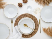 Draufsicht auf ein elegant gedecktes Tischarrangement mit weißen Tellern mit goldfarbenem Rand, goldfarbenem Besteck, geflochtenem Platzset, Weinglas und Wasserglas auf hellem Hintergrund mit Trockenblumen.