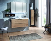 Moderne Badkombination mit Holzoptik, bestehend aus einem Waschtisch mit Schubladen, einem großen Spiegel und einem hohen Schrank, aus der Perspektive eines hellen Badezimmers mit Holzboden.