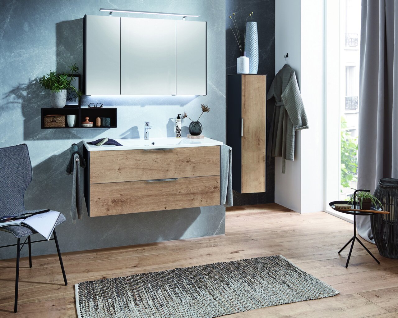 Moderne Badkombination mit Holzoptik, bestehend aus einem Waschtisch mit Schubladen, einem großen Spiegel und einem hohen Schrank, aus der Perspektive eines hellen Badezimmers mit Holzboden.