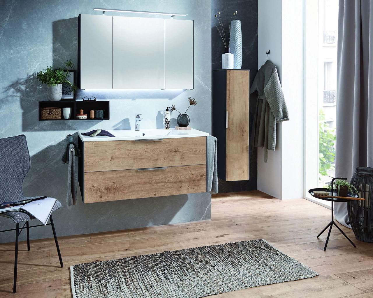 Moderne Badkombination mit Holzoptik, bestehend aus einem Waschtisch mit Schubladen, einem großen Spiegel und einem hohen Schrank, aus der Perspektive eines hellen Badezimmers mit Holzboden.