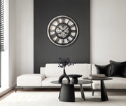 Moderne Wanduhr mit Zahnrädern in einem stilvollen Wohnzimmer, frontal an einer dunklen Wand über einem weißen Sofa montiert.