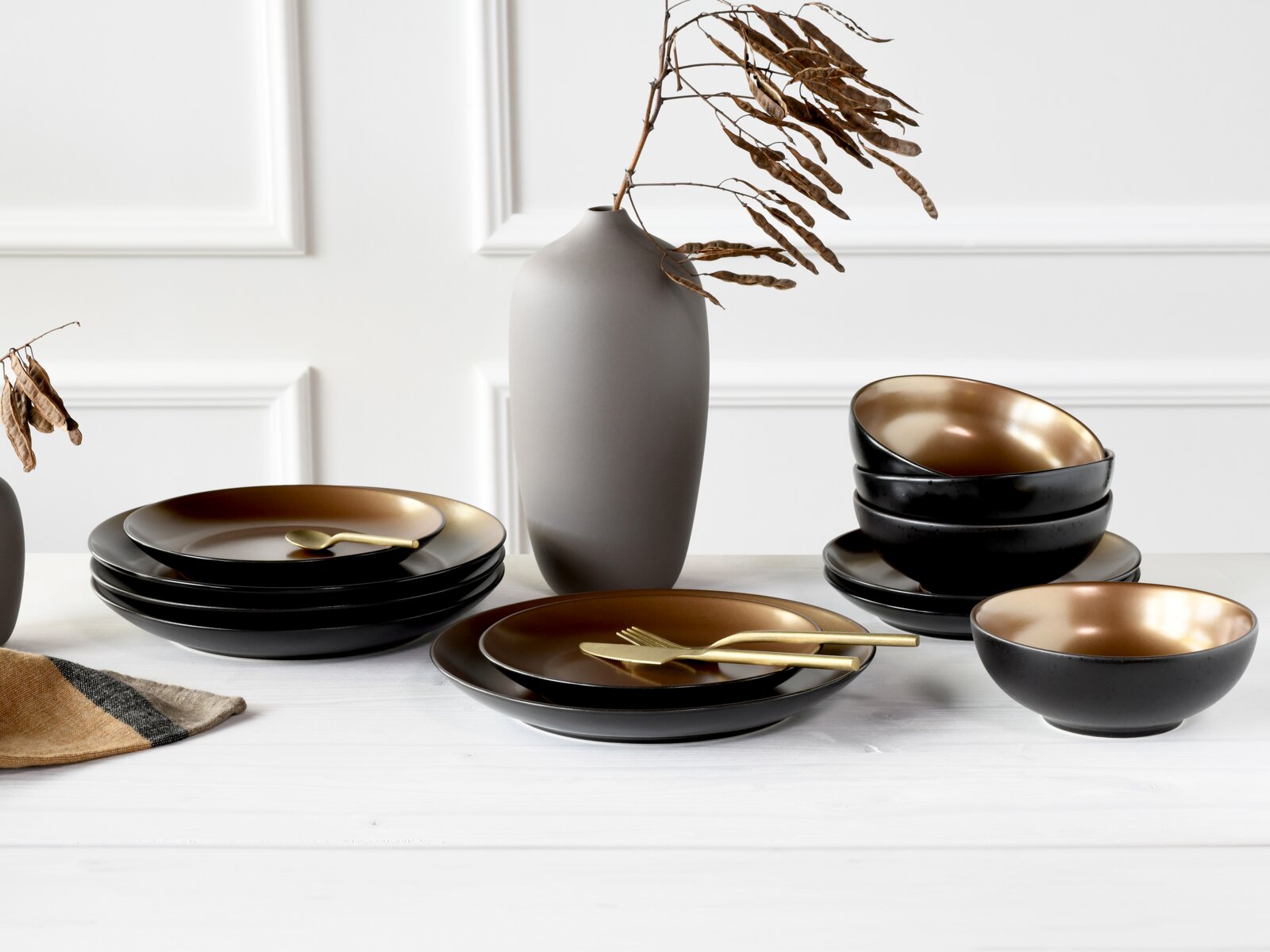Modernes Geschirr-Set mit schwarzen Tellern und Schalen mit goldfarbenem Innenfinish, goldfarbenem Besteck und grauer Vase auf weißem Tisch vor heller Wand, Perspektive: frontale leicht erhöhte Ansicht