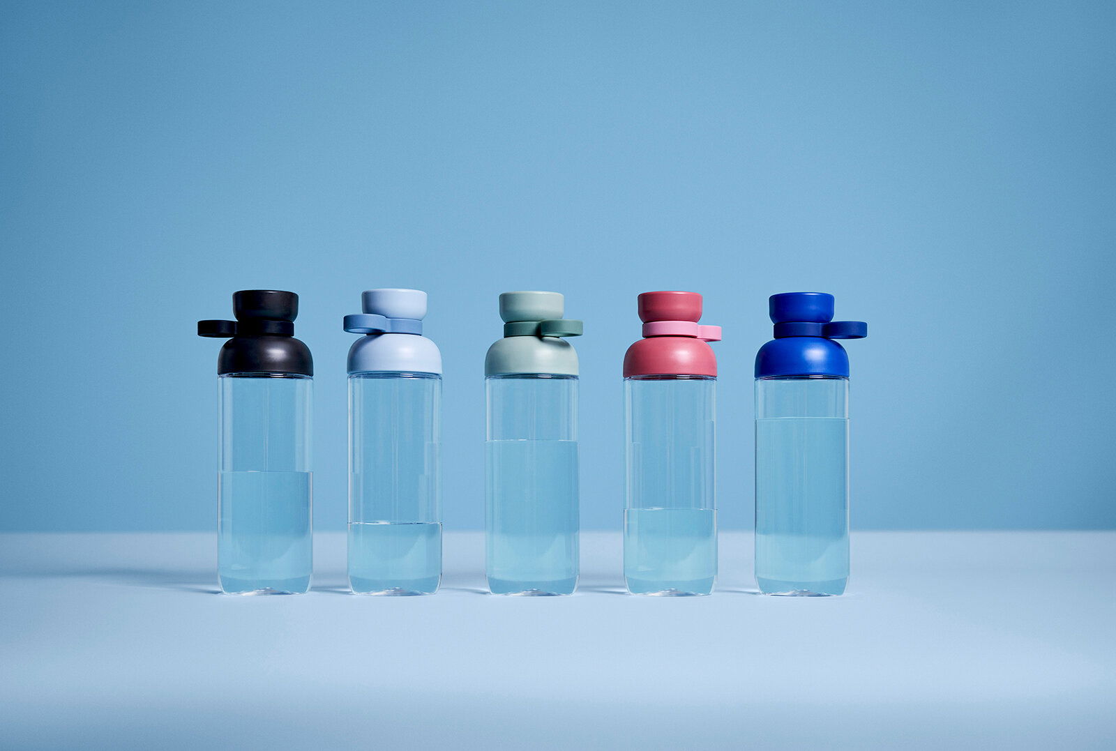 Fünf Trinkflaschen in verschiedenen Farben (schwarz, hellblau, grün, rosa, blau) stehen nebeneinander auf einer ebenen Fläche vor einem blauen Hintergrund. Die Flaschen sind aus transparentem Material und haben einen runden Deckel. Perspektive: Frontalans