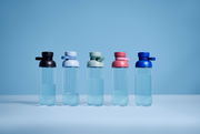 Fünf Trinkflaschen in verschiedenen Farben (schwarz, hellblau, grün, rosa, blau) stehen nebeneinander auf einer ebenen Fläche vor einem blauen Hintergrund. Die Flaschen sind aus transparentem Material und haben einen runden Deckel. Perspektive: Frontalans