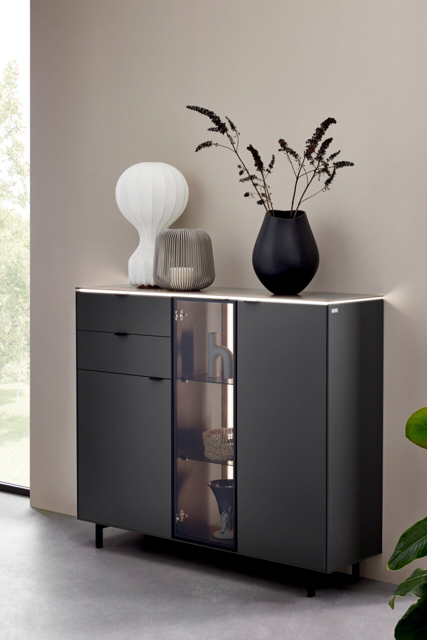 Modernes Highboard in dunklem Design mit Glasvitrine, dekoriert mit Vasen und Lampe, seitliche Perspektive.