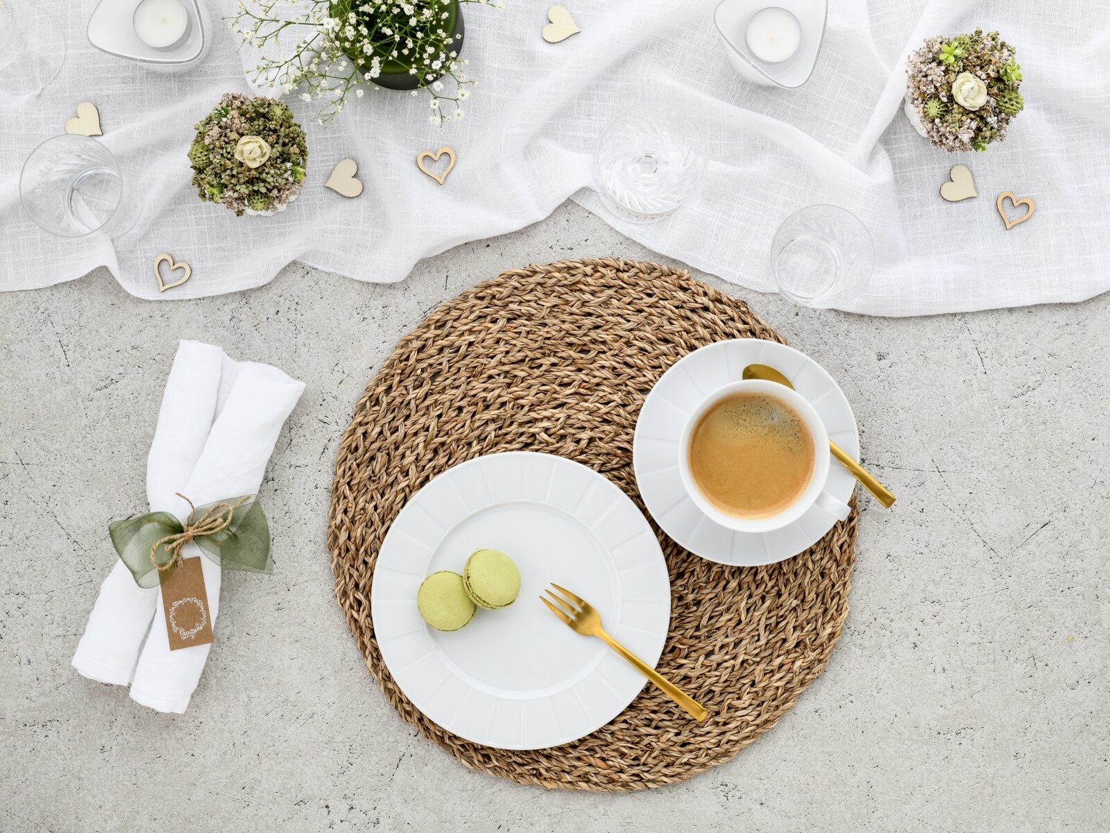 Draufsicht auf ein stilvolles Tischgedeck: weißes Geschirr auf rundem geflochtenem Platzset, Tasse Kaffee mit goldfarbenem Löffel, zwei grüne Macarons auf Teller mit goldfarbener Gabel, dazu helle Stoffdeko, Kerzen und kleine Herzornamente.