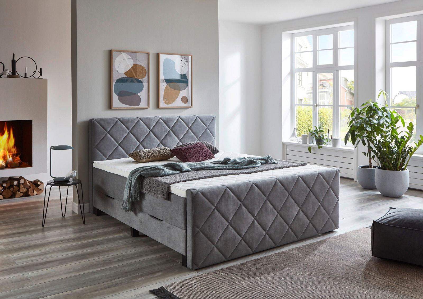 Graues Boxspringbett BX2200 CHESTER in einem modernen Schlafzimmer mit Kamin und großen Fenstern, seitliche Perspektive.