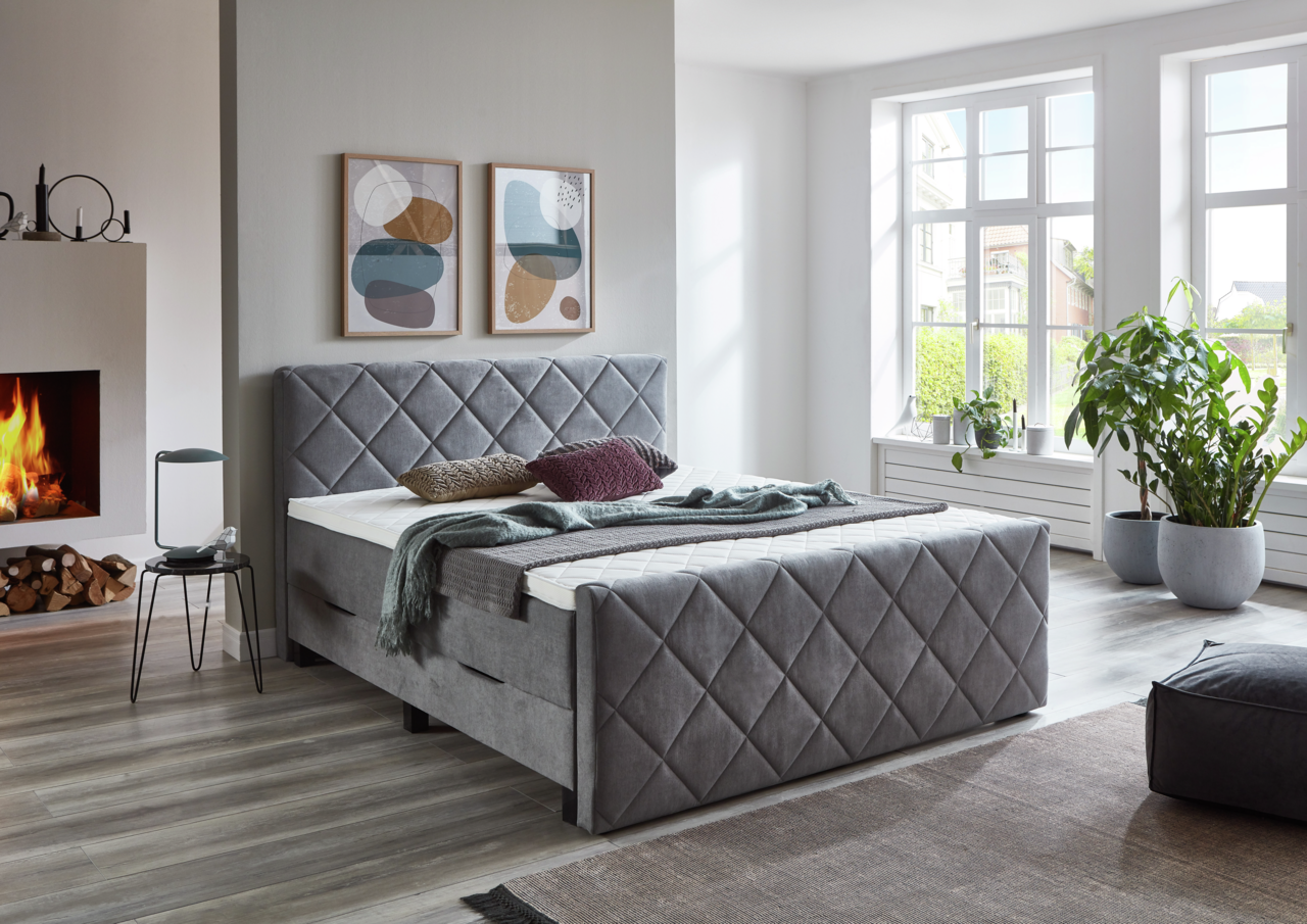 Z2 Boxbett BX2200 CHESTER Graues Boxspringbett BX2200 CHESTER in einem modernen Schlafzimmer mit Kamin und großen Fenstern, seitliche Perspektive.