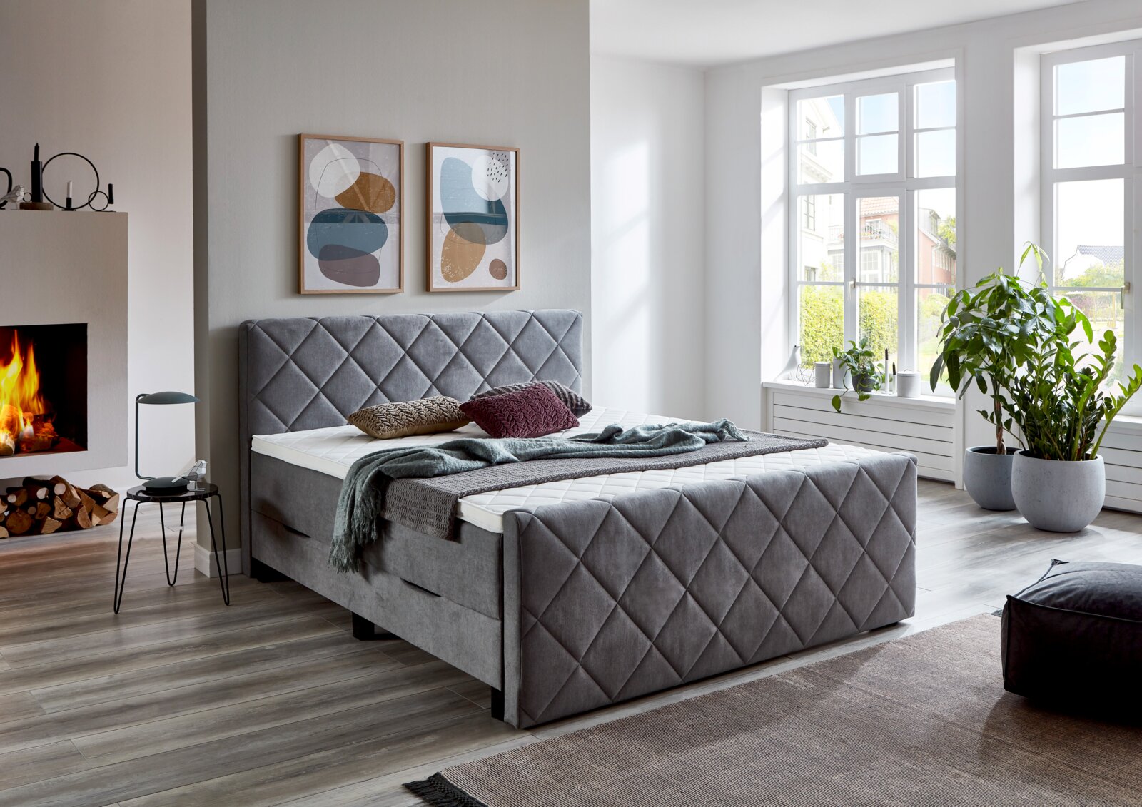 Graues Boxspringbett BX2200 CHESTER in einem modernen Schlafzimmer mit Kamin und großen Fenstern, seitliche Perspektive.