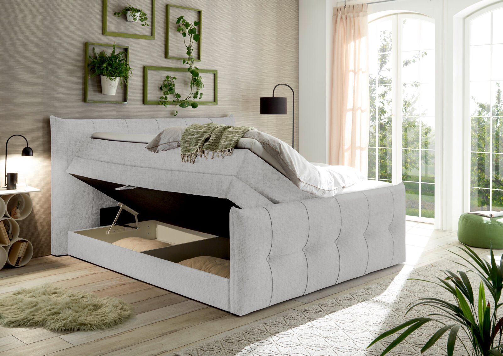 Boxspringbett mit Stauraum, seitliche Perspektive, in einem hellen Raum mit Pflanzen und großen Fenstern