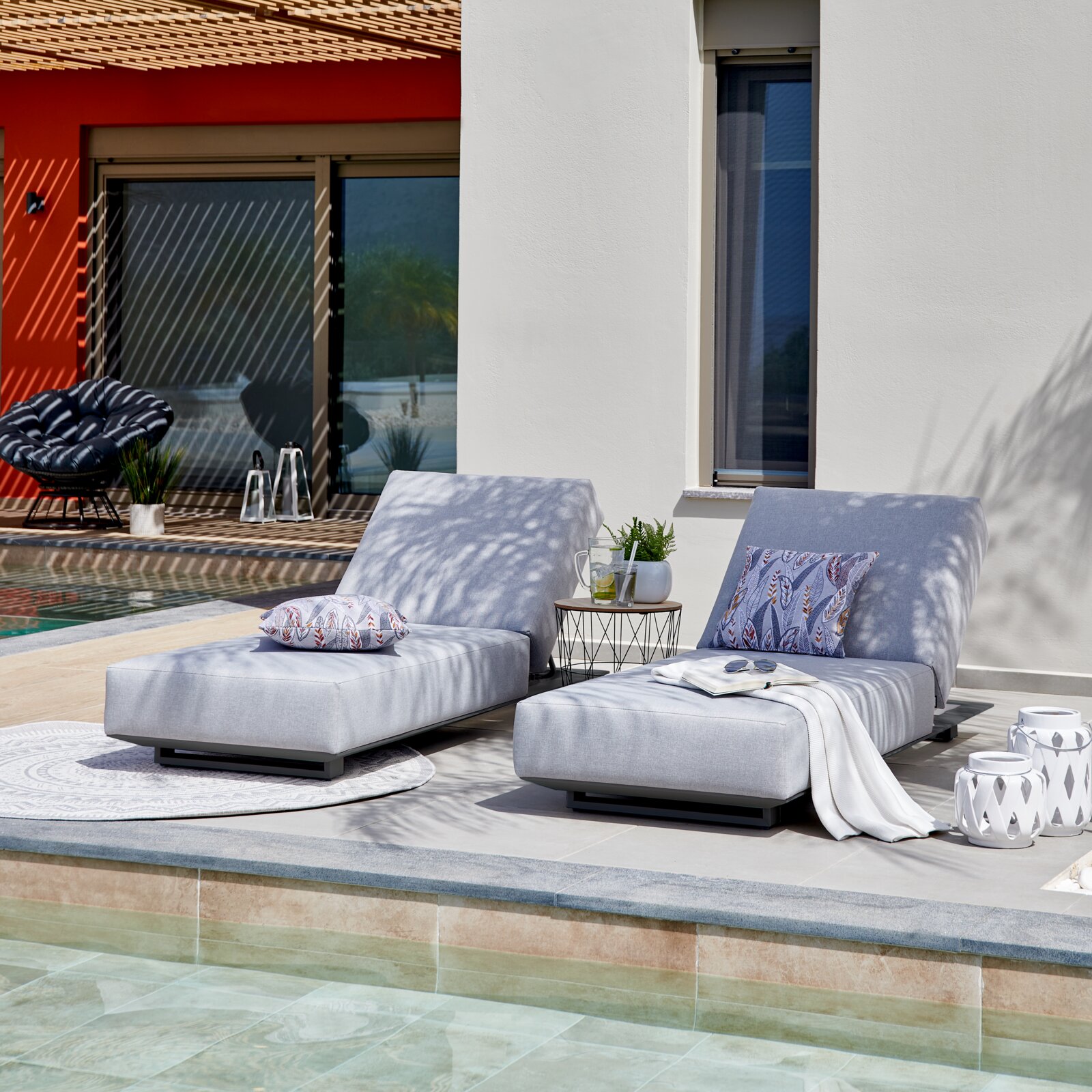 Twee moderne ligstoelen met grijze bekleding en decoratieve kussens, op een terras naast een zwembad, gefotografeerd vanuit een zijdelingse hoek.
