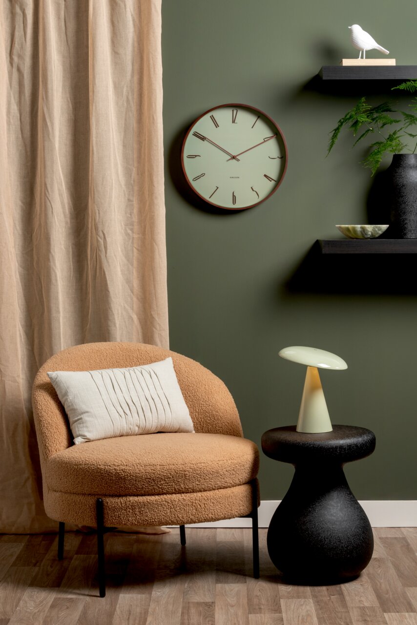 LED Akku Tischleuchte auf einem schwarzen Beistelltisch, seitliche Perspektive, neben einem beigen Sessel vor einer grünen Wand mit Uhr und Dekoration.