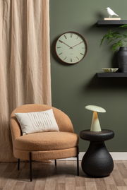 LED Akku Tischleuchte auf einem schwarzen Beistelltisch, seitliche Perspektive, neben einem beigen Sessel vor einer grünen Wand mit Uhr und Dekoration.