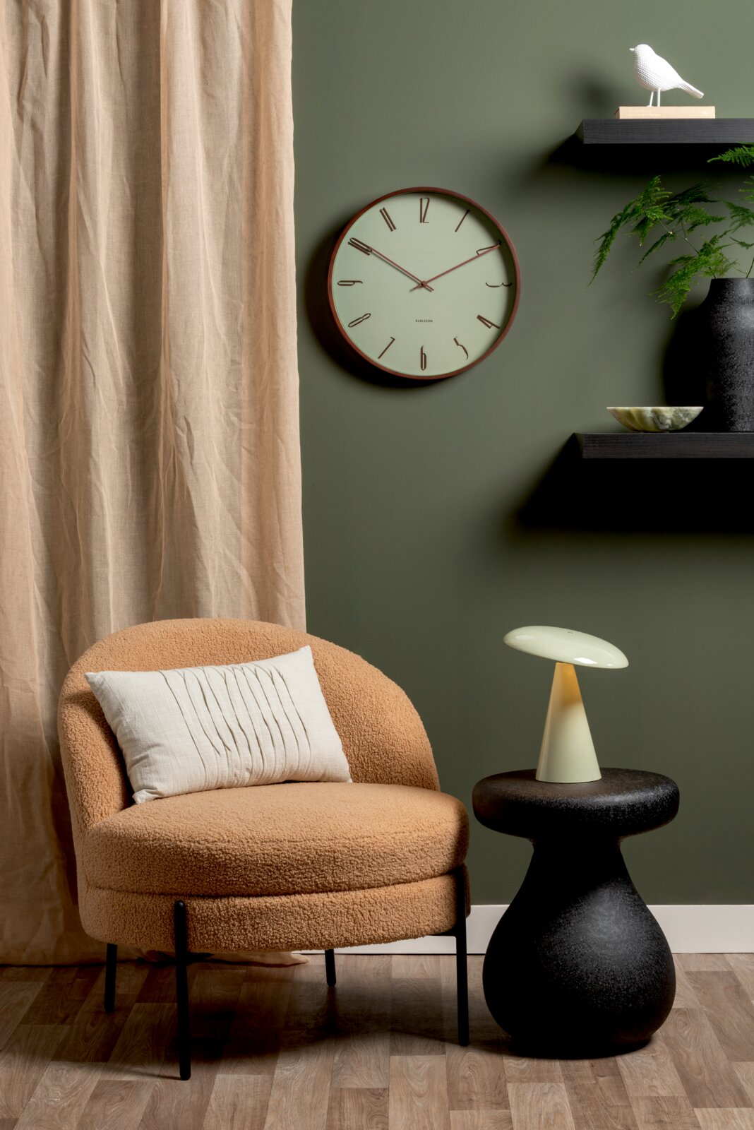 LED-tafellamp op een zwarte bijzettafel, gezien vanaf de zijkant, naast een beige fauteuil voor een groene muur met een klok en decoratie.