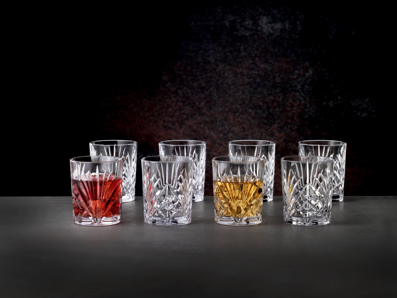 Set aus acht Kristallgläsern mit elegantem Schliffmuster, gefüllt mit verschiedenen Getränken, auf einer dunklen Oberfläche vor schwarzem Hintergrund, aus frontaler Perspektive.