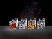 Set aus acht Kristallgläsern mit elegantem Schliffmuster, gefüllt mit verschiedenen Getränken, auf einer dunklen Oberfläche vor schwarzem Hintergrund, aus frontaler Perspektive.
