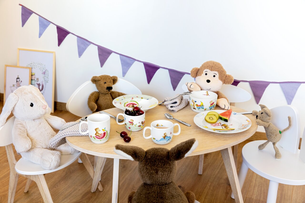 Villeroy & Boch Kinderbesteck 4 tlg. HAPPY AS A BEAR Kinderbesteck-Set auf einem Tisch mit Plüschtieren, Teller und Tassen mit bunten Motiven, seitliche Perspektive