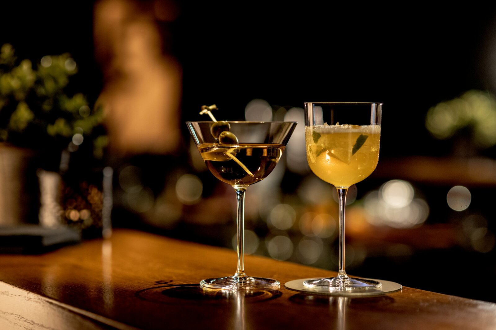 Zwei Cocktails in eleganten Stielgläsern auf einer Bar-Theke, links ein klarer Drink im Coupéglas mit Olivenspieß, rechts ein gelber Drink im Kelchglas mit Minze und Eis, frontal aus Augenhöhe fotografiert mit unscharfem Bar-Hintergrund.