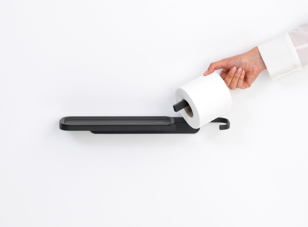 Moderner Toilettenpapierhalter mit Ablage in seitlicher Perspektive, Hand legt eine Toilettenpapierrolle auf den Halter.
