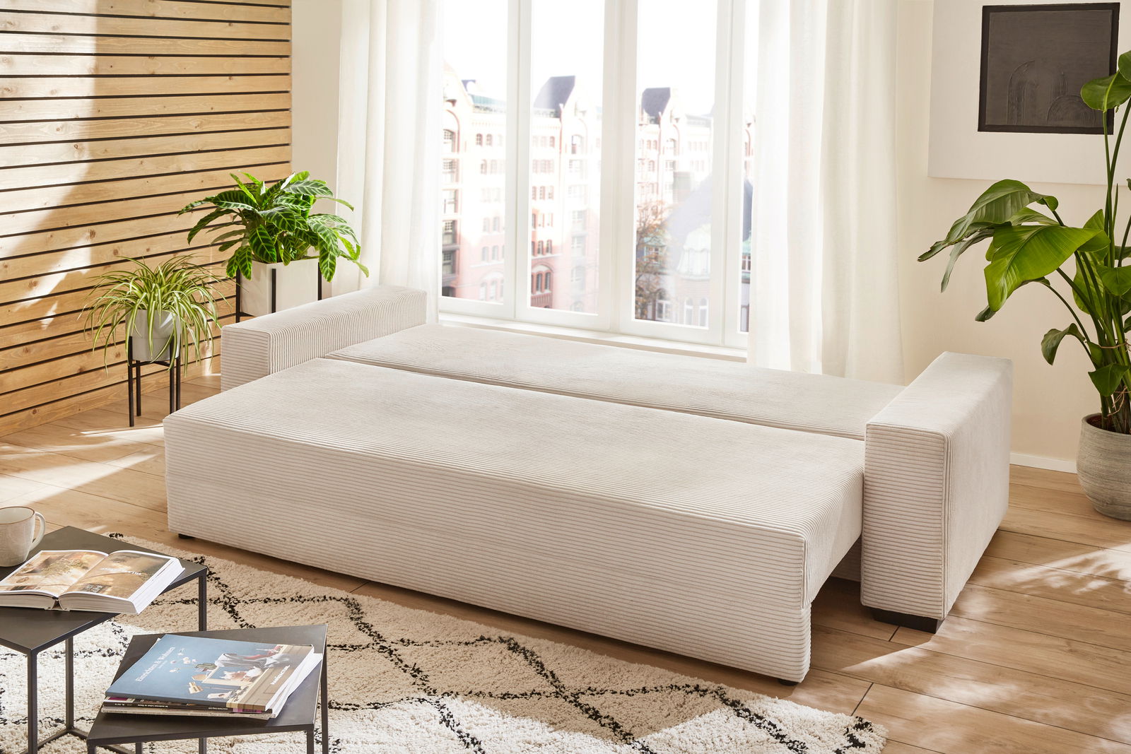 Funktionssofa in heller Farbe, ausgeklappt als Bett, in einem modernen Wohnzimmer mit Pflanzen und Holzwand, seitliche Perspektive