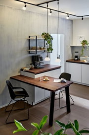 Moderne Küche mit Schienenspot-Beleuchtung von oben, die eine gemütliche Atmosphäre schafft. Perspektive von der Seite auf den Esstisch und die Küchenzeile.