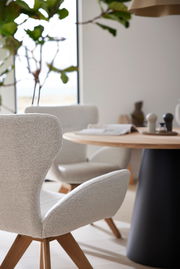 Moderner Armlehnstuhl mit hellem Stoffbezug und Holzbeinen, im Vordergrund eines stilvoll eingerichteten Essbereichs mit rundem Tisch. Seitliche Perspektive.