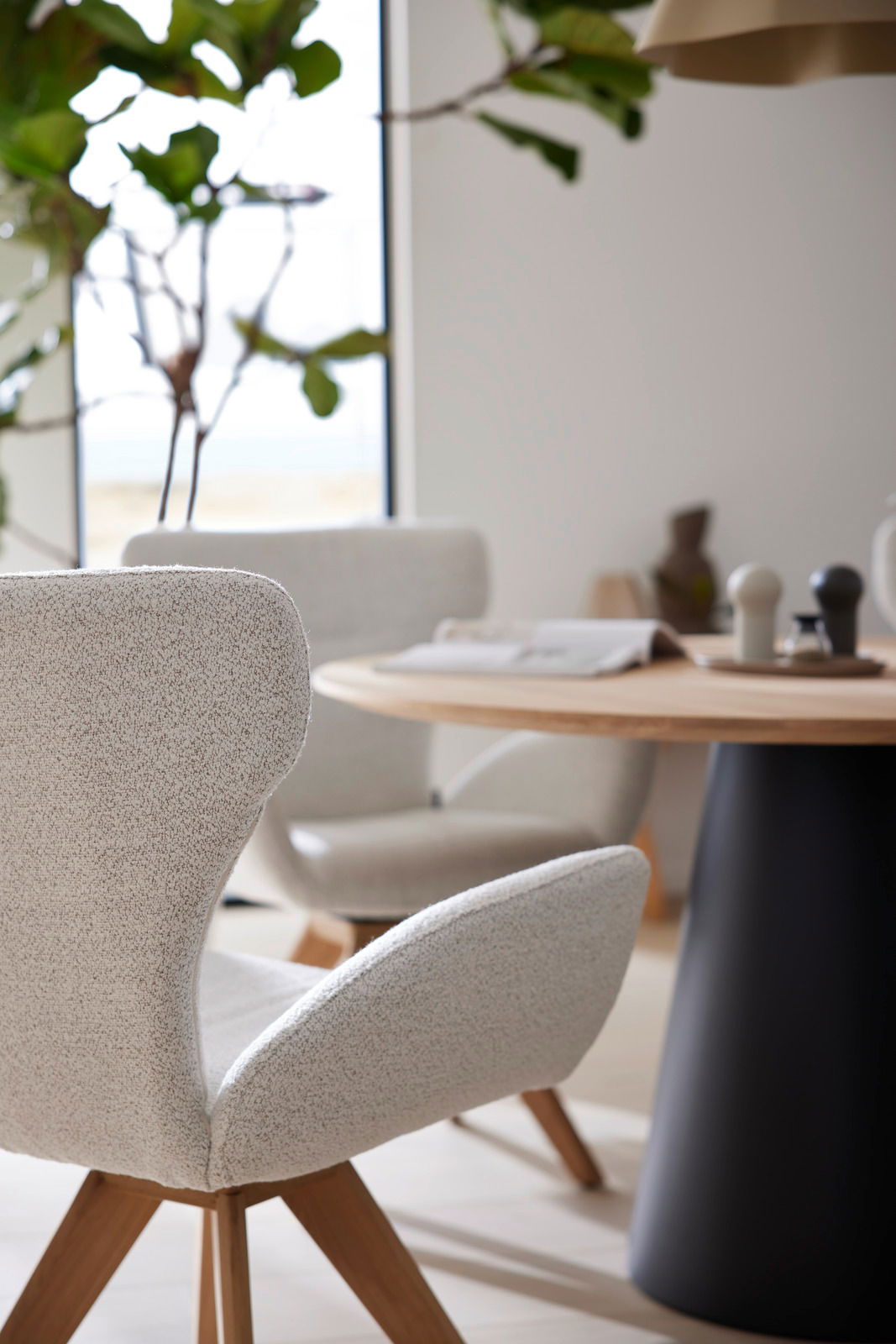 Moderner Armlehnstuhl mit hellem Stoffbezug und Holzbeinen, im Vordergrund eines stilvoll eingerichteten Essbereichs mit rundem Tisch. Seitliche Perspektive.