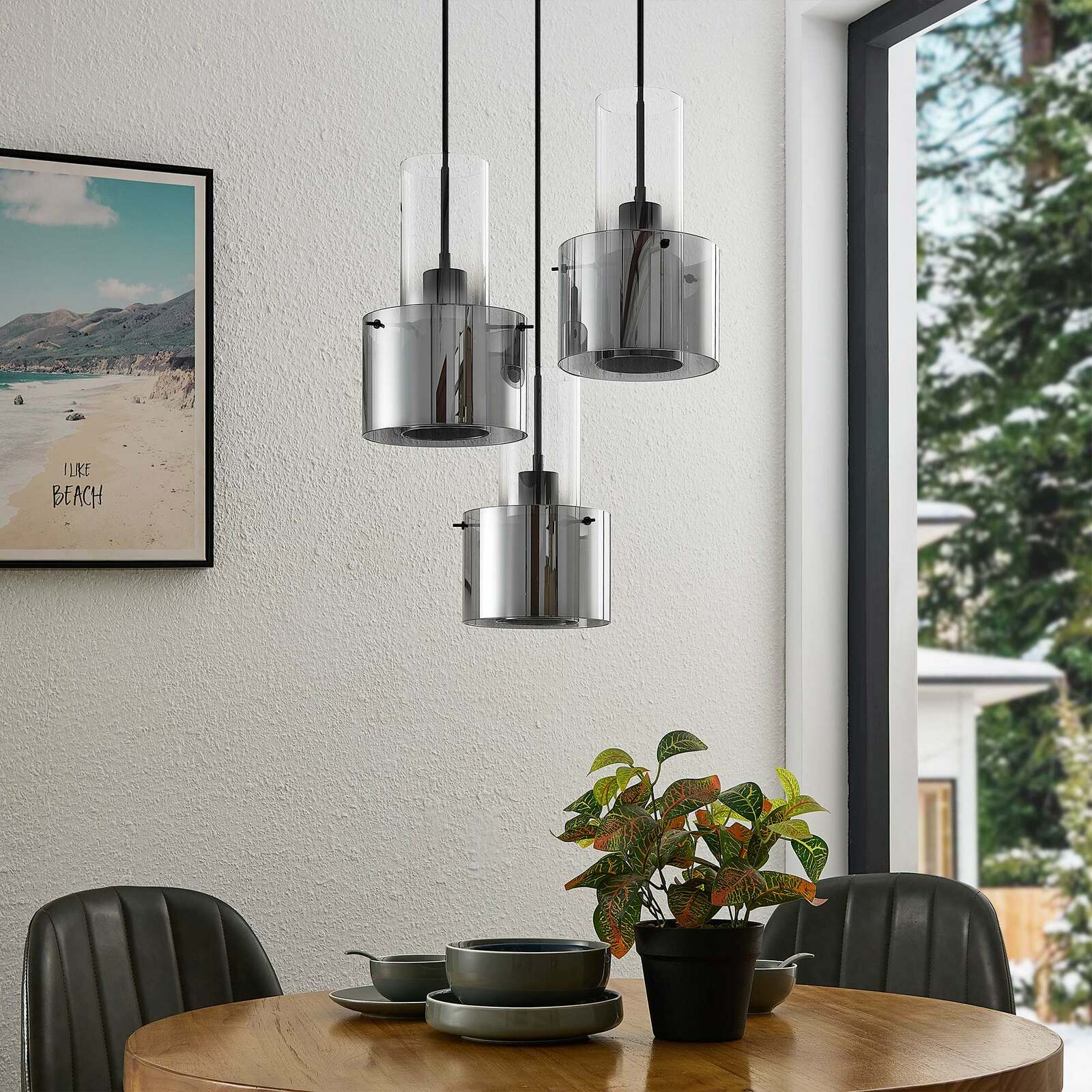 Moderne Hängeleuchte mit drei zylindrischen, rauchgrauen Glasschirmen, hängend über einem Esstisch in einem stilvoll eingerichteten Raum, seitliche Perspektive.