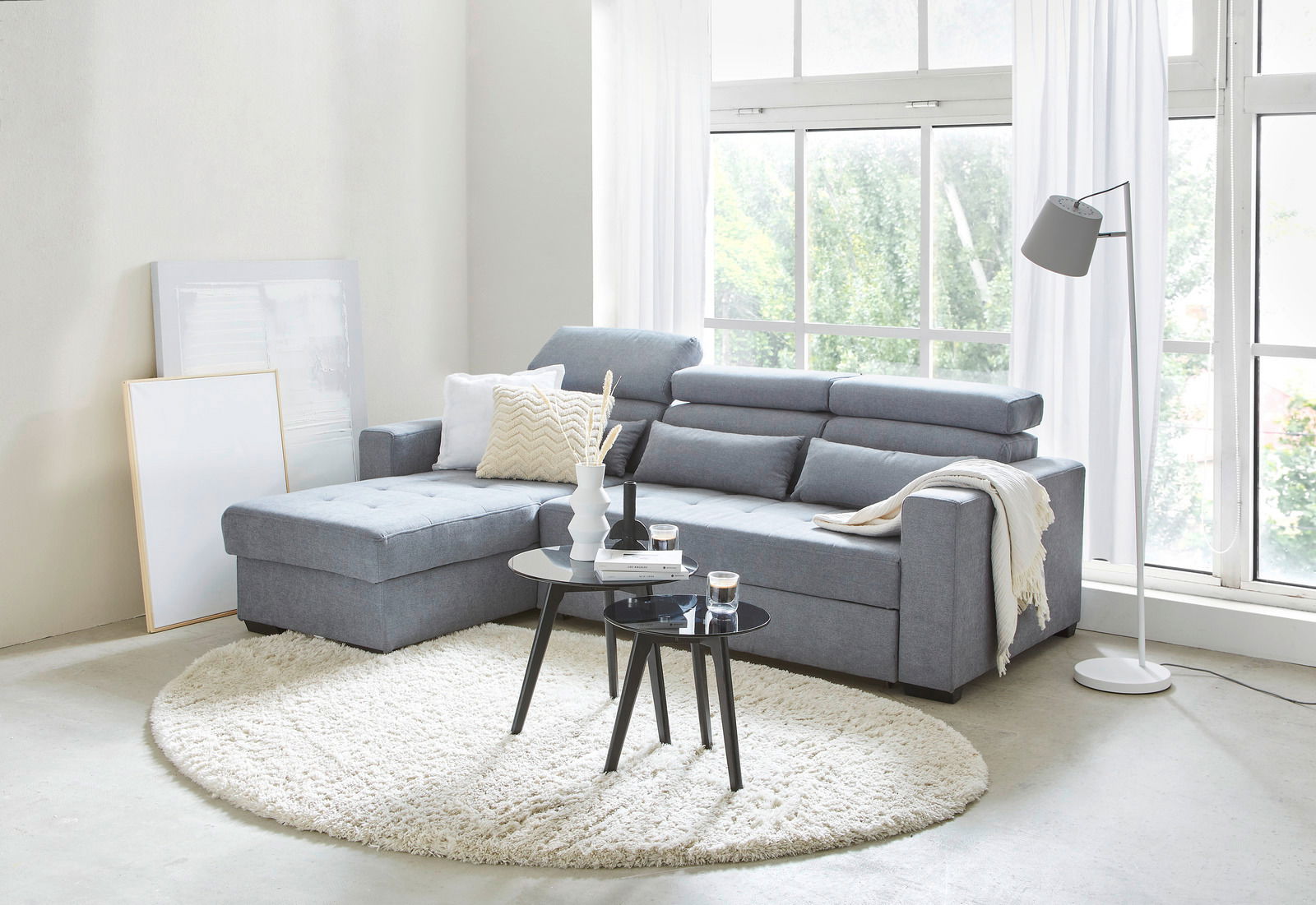 Graues Ecksofa in modernem Wohnzimmer mit großen Fenstern, runder Teppich und zwei Couchtischen, seitliche Perspektive.