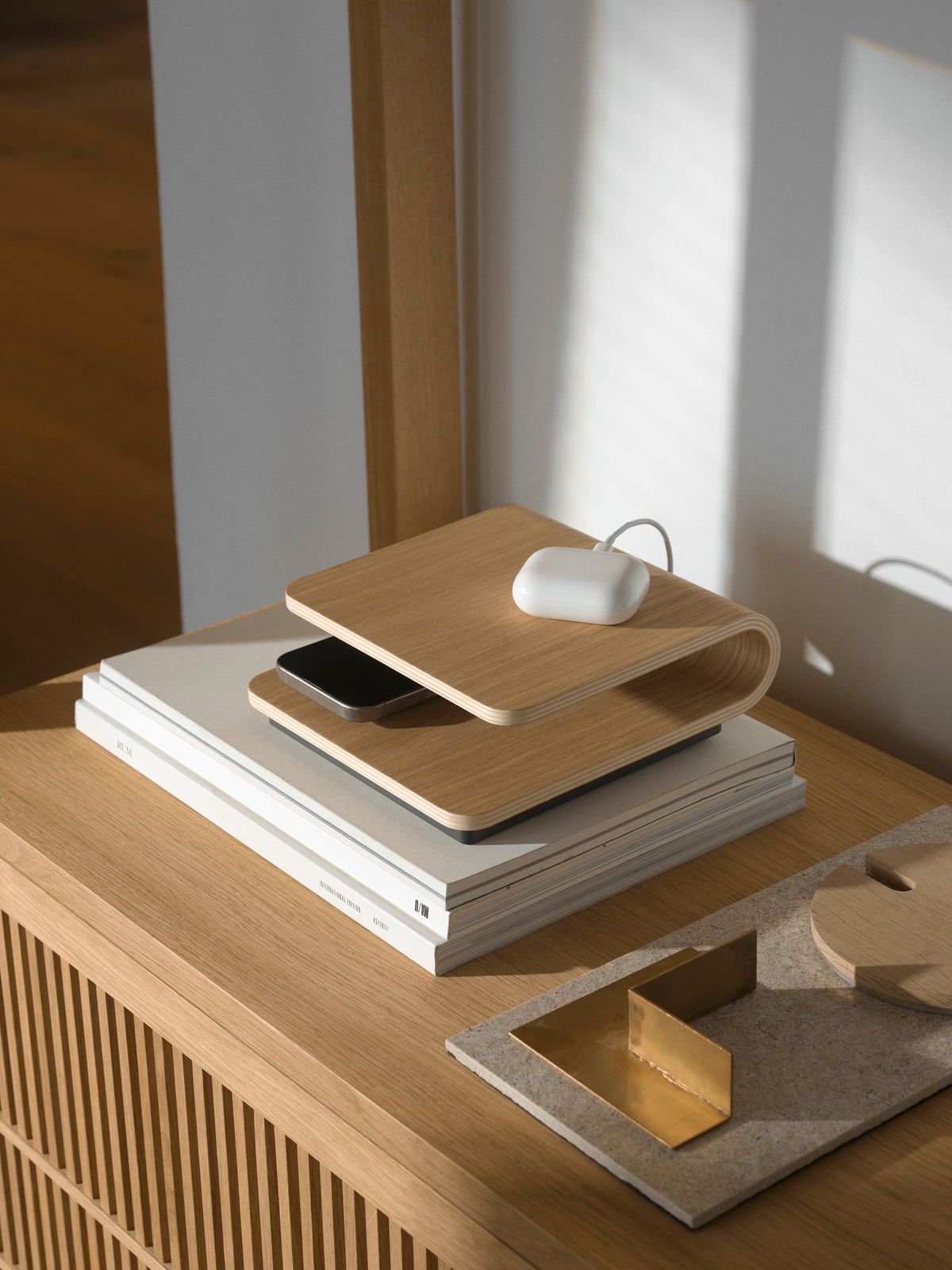 Stelton Draadloze oplader RO Kabelloses Ladegerät aus Holz in seitlicher Perspektive auf einem Tisch, mit einem Smartphone und einem weißen Ladecase darauf.