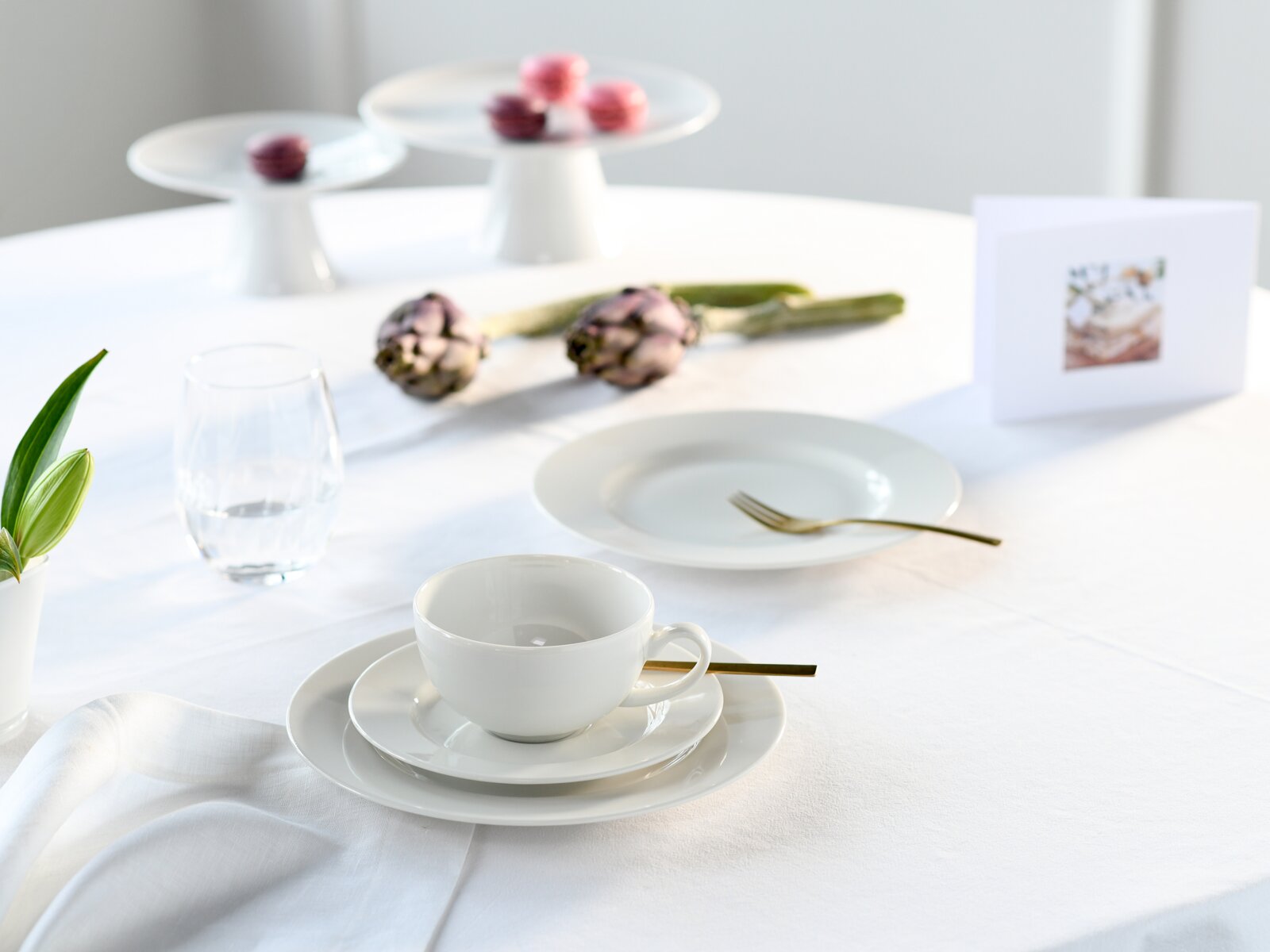 Schlichtes weißes Porzellan-Kaffeegedeck mit Tasse, Untertasse und Teller auf weißer Tischdecke, daneben goldfarbenes Besteck und Wasserglas; Perspektive: leicht erhöhte Frontansicht mit Fokus auf die Tasse im Vordergrund.