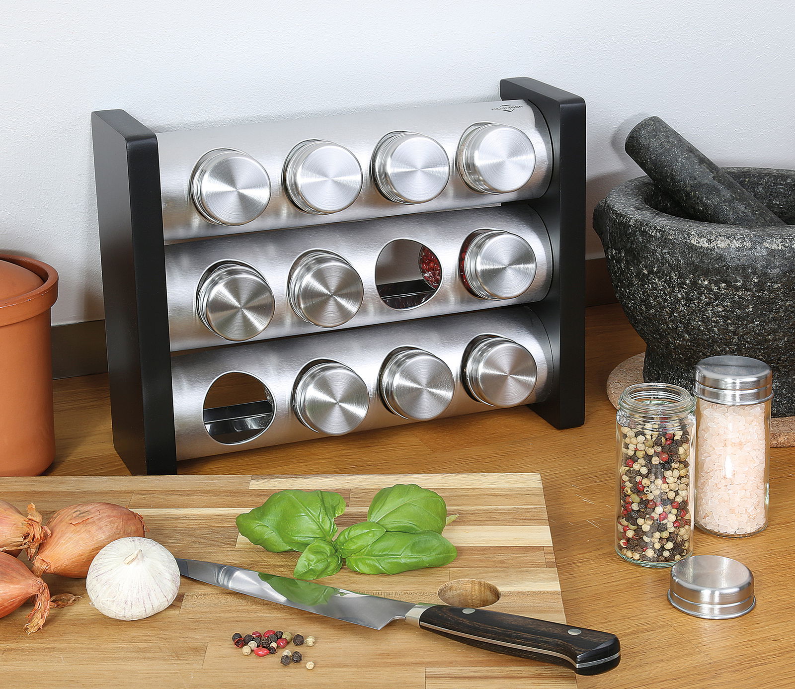 Gewürzständer mit 12 Gläsern aus Edelstahl, frontal auf einer Küchenarbeitsplatte mit Gewürzen und Küchenutensilien.