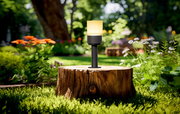 Ein modernes Akku-Windlicht auf einem Baumstumpf im Garten, umgeben von bunten Blumen und grünem Laub, aus der Perspektive von vorne betrachtet.