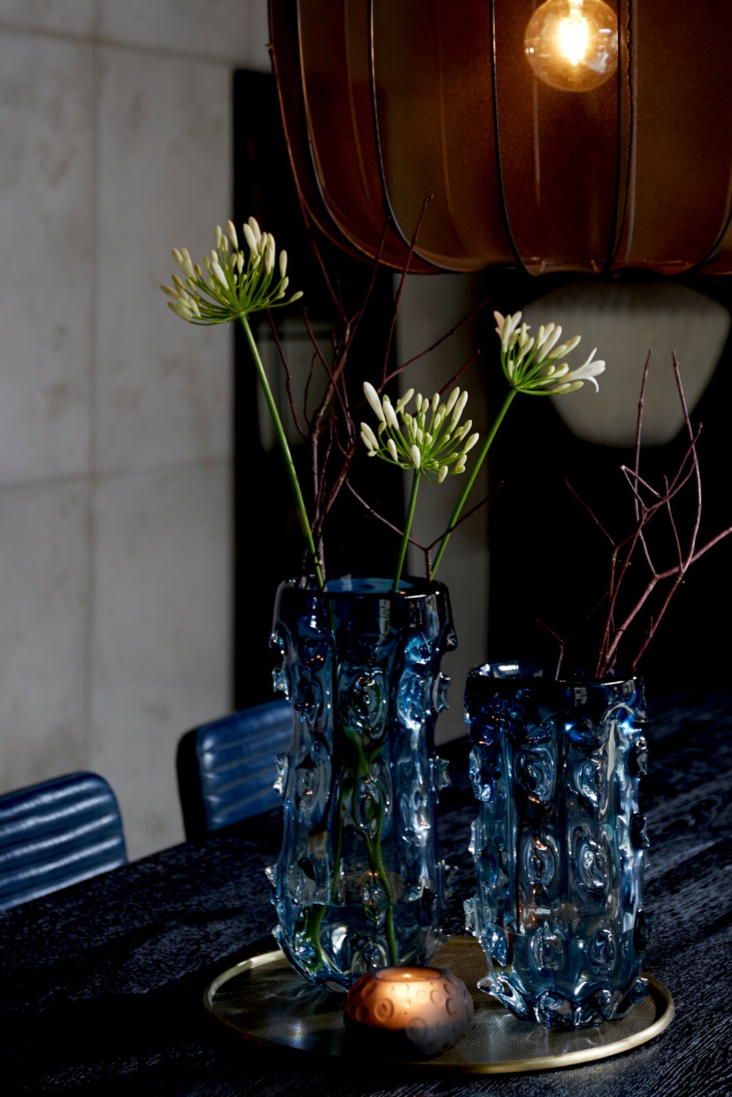Twee sculpturale blauwe glazen vazen met bloemen en takken op een ronde schaal, sfeervol van voren en licht van boven gefotografeerd onder een warme hanglamp.