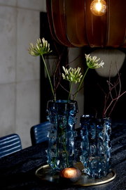 Twee sculpturale blauwe glazen vazen met bloemen en takken op een ronde schaal, sfeervol van voren en licht van boven gefotografeerd onder een warme hanglamp.