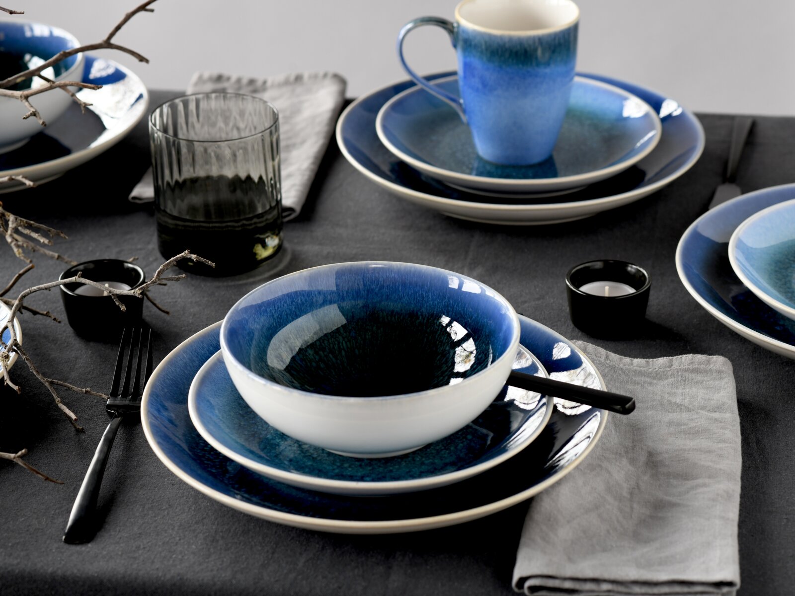 Schlicht gedeckter Tisch mit blau glasiertem Geschirr-Set aus Teller, Schale und Tasse, dazu Besteck und Glas, fotografiert in schräger Draufsicht.