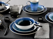 Schlicht gedeckter Tisch mit blau glasiertem Geschirr-Set aus Teller, Schale und Tasse, dazu Besteck und Glas, fotografiert in schräger Draufsicht.