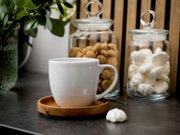 Weiße Keramiktasse auf Holzuntertasse mit Löffel auf dunkler Arbeitsplatte, im Hintergrund zwei Glasvorratsdosen mit Keksen und Baiser sowie grüne Pflanze; Perspektive: frontale Nahaufnahme auf Augenhöhe.