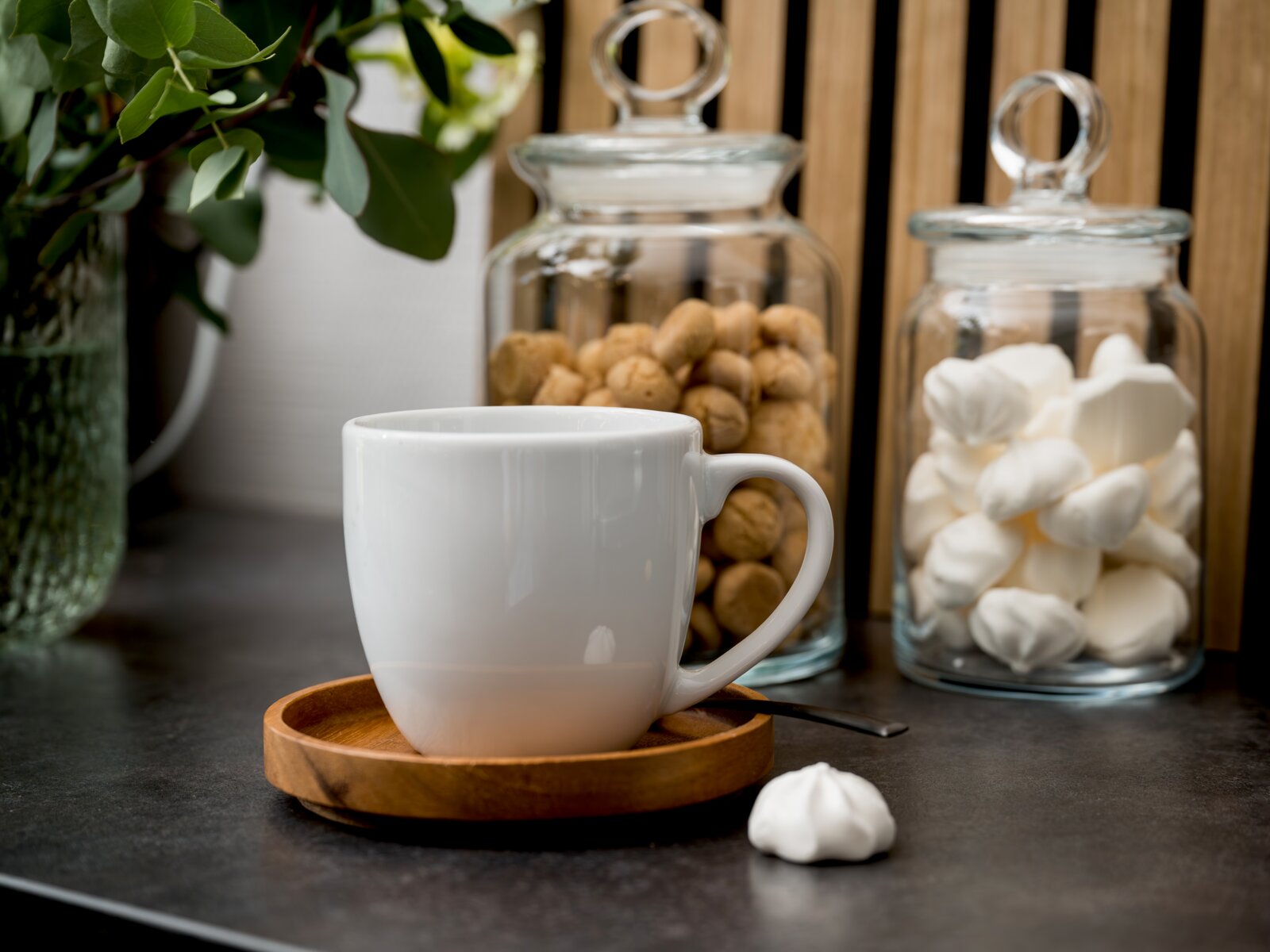Weiße Keramiktasse auf Holzuntertasse mit Löffel auf dunkler Arbeitsplatte, im Hintergrund zwei Glasvorratsdosen mit Keksen und Baiser sowie grüne Pflanze; Perspektive: frontale Nahaufnahme auf Augenhöhe.