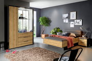 Modernes Schlafzimmer mit Bett, Nachttisch und Kleiderschrank in Holzoptik mit schwarzen Akzenten; Kleiderschrank mit Glasfront und Schubladen, Bett mit gepolstertem Kopfteil – Perspektive: frontale Raumansicht in leichter Schrägansicht von links.