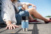 Thermobecher 0,3L in hellblau mit Edelstahl- und schwarzem Deckel, im Vordergrund auf dem Boden stehend, mit unscharfer Person im Hintergrund.