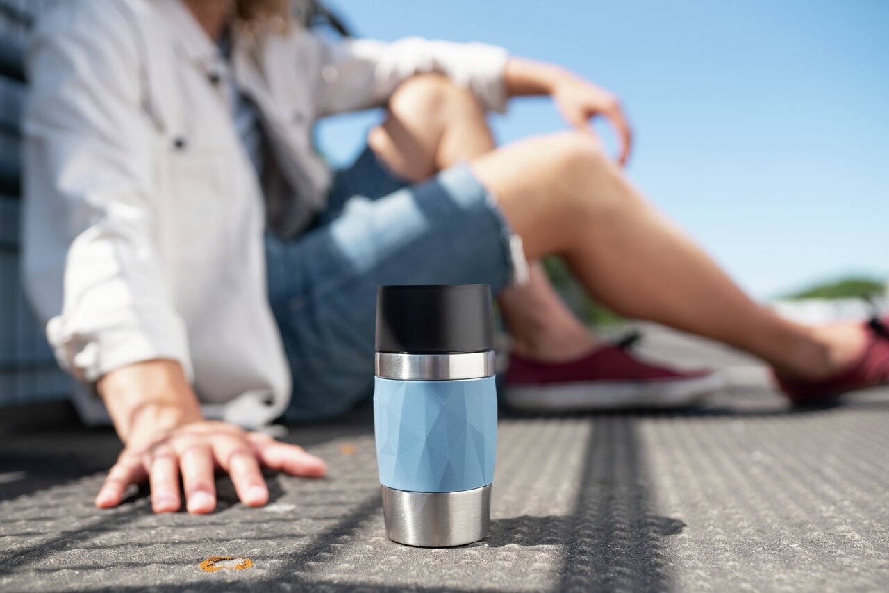 Thermobecher 0,3L in hellblau mit Edelstahl- und schwarzem Deckel, im Vordergrund auf dem Boden stehend, mit unscharfer Person im Hintergrund.