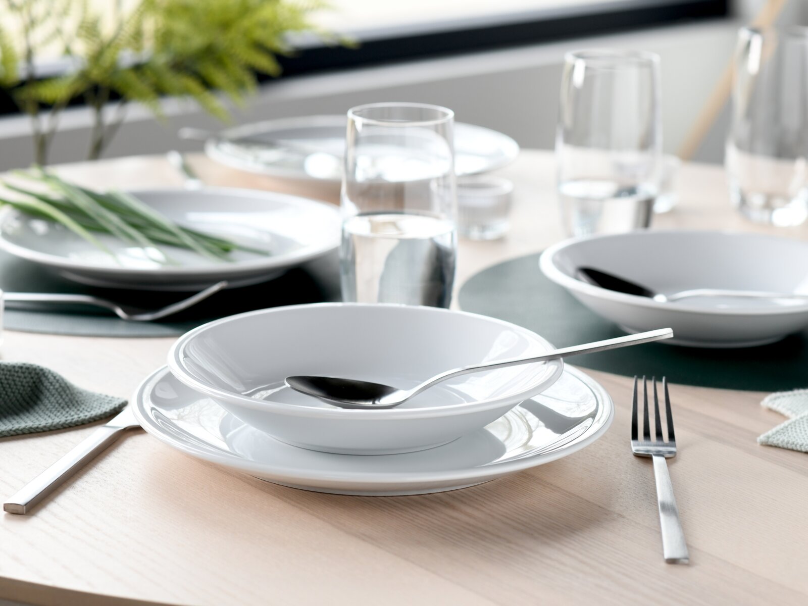 Modern gedeckter Esstisch mit weißem Geschirr (tiefer Teller/Schale auf Speiseteller), Edelstahlbesteck und Wassergläsern; Perspektive: leicht erhöhte, schräge Nahaufnahme von vorn.
