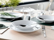 Modern gedeckter Esstisch mit weißem Geschirr (tiefer Teller/Schale auf Speiseteller), Edelstahlbesteck und Wassergläsern; Perspektive: leicht erhöhte, schräge Nahaufnahme von vorn.