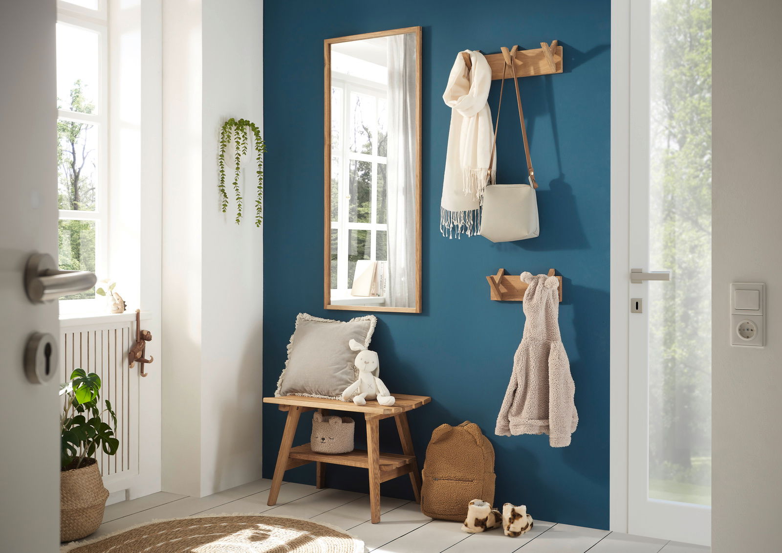 Eingangsbereich mit einer Garderobenbank aus Holz, einem Spiegel und Hakenleisten an einer blauen Wand. Perspektive von der Tür aus.