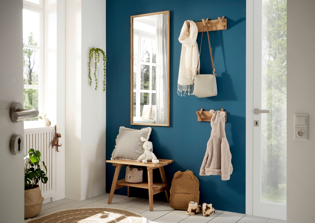Eingangsbereich mit einer Garderobenbank aus Holz, einem Spiegel und Hakenleisten an einer blauen Wand. Perspektive von der Tür aus.
