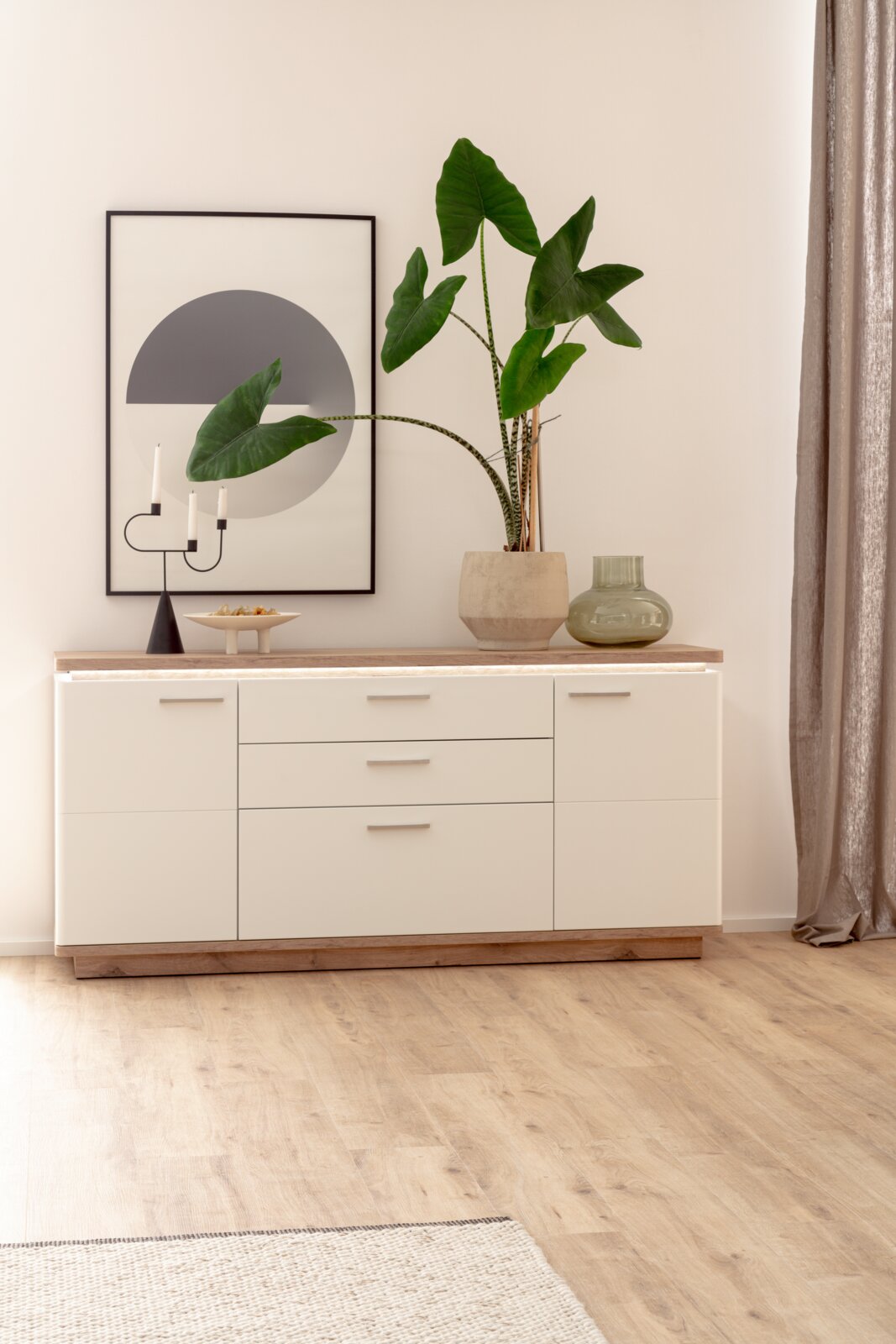Frontale Perspektive: moderne weiße Kommode mit Holzplatte und Holzsockel, dekoriert mit großer grüner Zimmerpflanze im Topf, Glasvase, Kerzenhalter und gerahmtem Wandbild im minimalistischen Wohnraum.